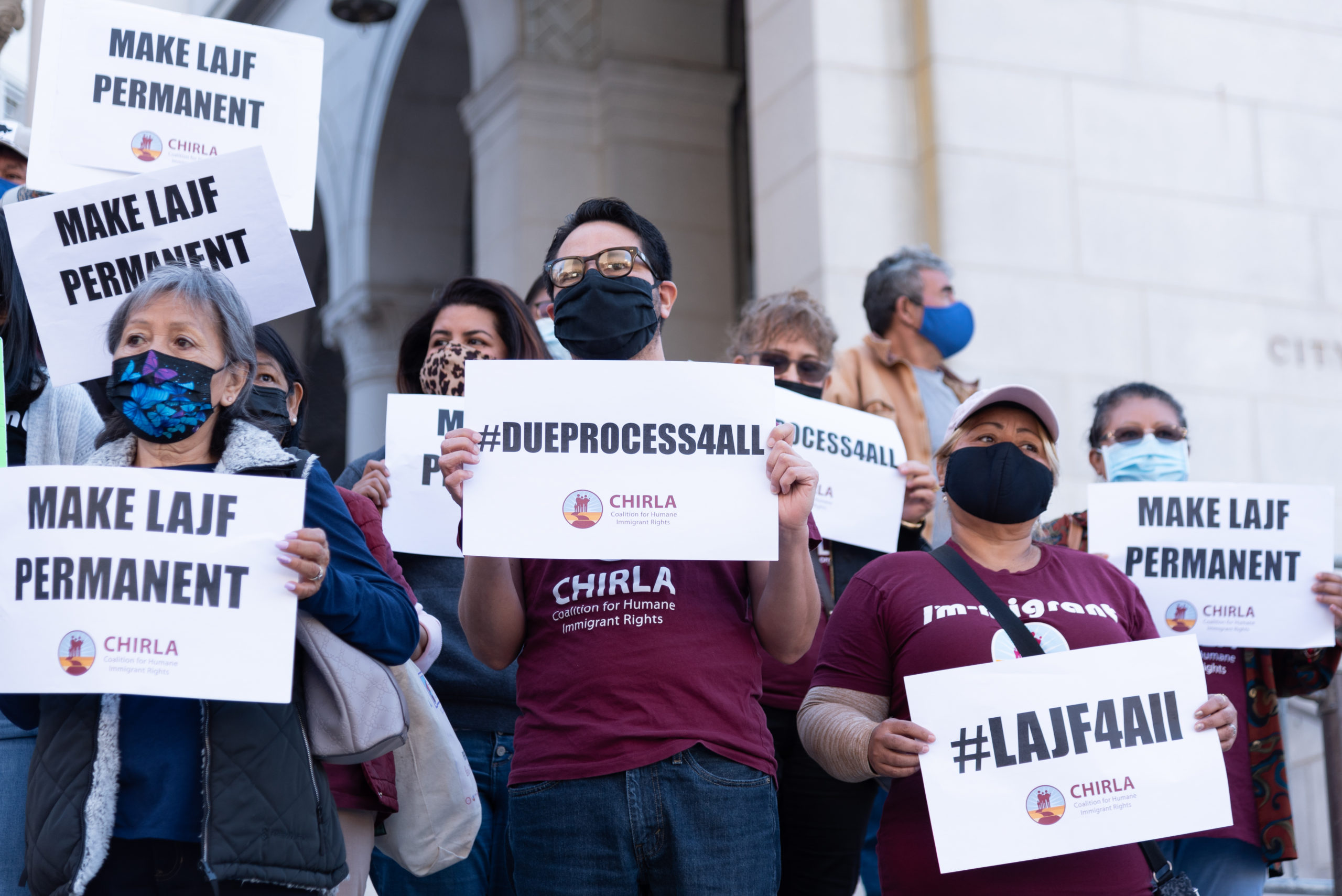 Protester wearing a red CHIRLA shirt and black mask holding a sign that says #DueProcess4All in front of a crowd of other protestors.